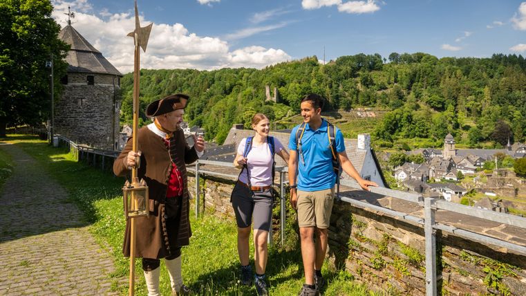 Ein Stadtführer in historischer Kleidung spricht mit zwei Wanderern auf einem malerischen Weg. Im Hintergrund sind grüne Hügel und ein Teil der Stadt sichtbar.