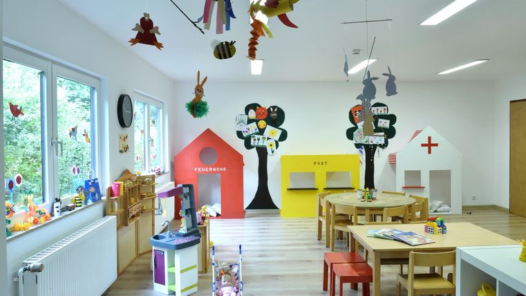 Ein heller und farbenfroher Raum mit Spielbereichen für Kinder. Es gibt bunte Spielhäuser, Tische und Spielsachen, die eine einladende Lernumgebung schaffen.