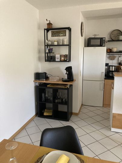 A modern kitchen with a small table and black chairs. In the background, there is a refrigerator, a microwave, and a coffee pot on a black shelf.