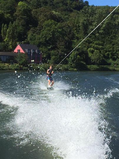 A man is standing on a wakeboard and is being towed across the water by a boat. In the background, there are trees and a pink house visible.