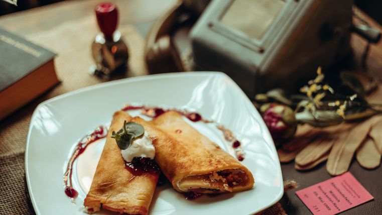 A plate with two filled crepes, garnished with cream and a fruity sauce. In the background, decorative items and a few books are visible.