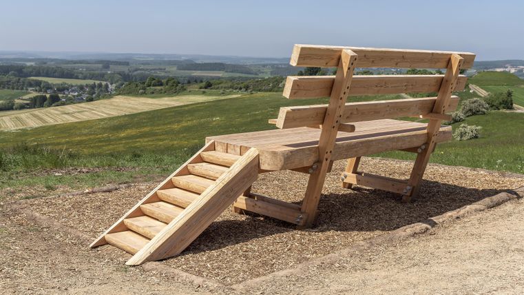 An oversized wooden bench with stairs stands on a hill overlooking fields and forests under a blue sky.
