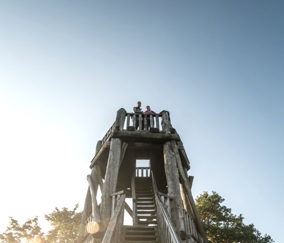 Holzaussichtsturm an der Dietzenley mit zwei Personen oben, umgeben von Bäumen und blauem Himmel., © Eifel Tourismus GmbH, Dominik Ketz