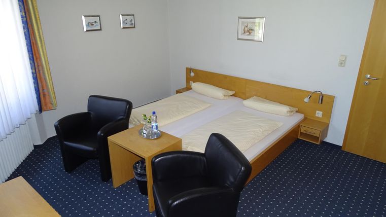 Ein einfaches Hotelzimmer mit einem Doppelbett und zwei schwarzen Stühlen. Der Raum hat einen neutralen Farbton und ein Fenster, das Tageslicht hereinlässt.