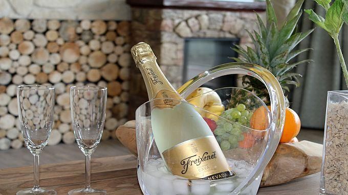 Eine Flasche Sekt in einem Kühler auf Eis, umgeben von frischen Früchten und zwei Gläsern. Der Hintergrund hat eine gemütliche Atmosphäre mit Holzscheiten.