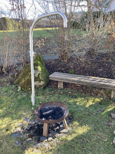 Ein Grillplatz im Garten mit einem metallenen Grillhaken und einer runden Feuerstelle. Im Hintergrund ist eine Bank und einiges an Grün zu sehen.
