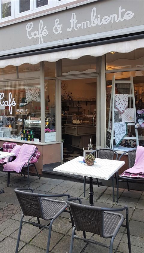 Ein einladendes Café mit Tischen und Stühlen im Außenbereich. Die Fenster zeigen eine gemütliche Inneneinrichtung.