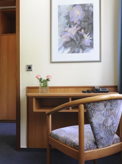 Ein heller Raum mit einem Holzstuhl und einem Schreibtisch. An der Wand hängt ein Blumenbild und auf dem Tisch steht eine kleine Blumenvase.