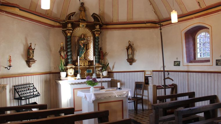 A small chapel with an altar, surrounded by wooden benches. Religious statues and pictures hang on the walls.