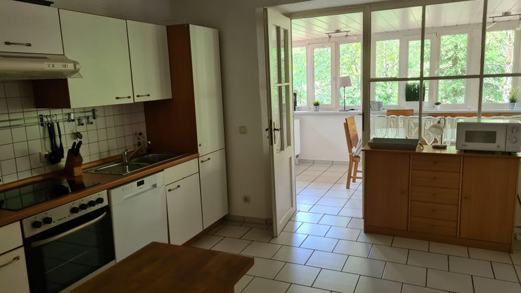 Eine helle Küche mit weißen Schränken und einer Holzarbeitsplatte. Durch große Fenster scheint viel Licht herein, und ein Tisch befindet sich im angrenzenden Raum.