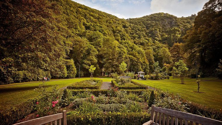 Ein schöner Garten mit gepflegten Blumenbeeten und Rasenflächen. Im Hintergrund sind bewaldete Hügel zu sehen.