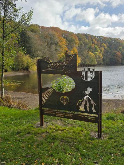 Een houten bord aan de oever van een meer, omringd door kleurrijk herfstblad. Op de achtergrond zijn bomen en een rustig water te zien.