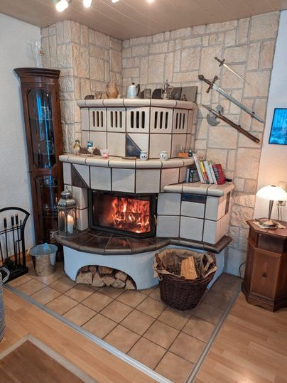 A cozy tiled stove with a flaming fire in a bright room. Next to it are a wood basket and furniture that create a warm atmosphere.
