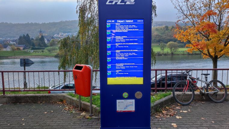 Ein Fahrkartenautomat mit Informationen über Zugverbindungen steht an einem ruhigen Flussufer. Im Hintergrund sind Bäume und Fahrräder zu sehen.
