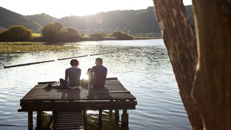 Zwei Personen sitzen auf einem Holzsteg am Wasser und genießen den Sonnenuntergang. Im Hintergrund sind sanfte Hügel und grüne Vegetation sichtbar.