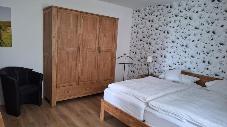 Een eenvoudig slaapkamer met een tweepersoonsbed en een grote houten kast. Naast het bed staat een zwarte fauteuil en een wand met een bloemenpatroon.