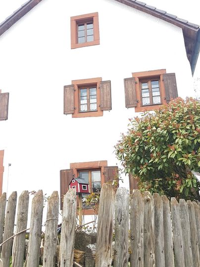 A charming white house with brown shutters. In the foreground, there is a simple wooden fence and a slightly overgrown garden.