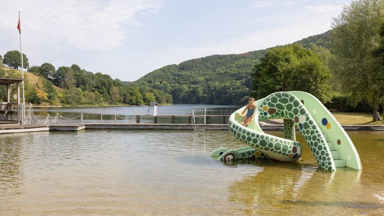 Ein Wasserspielplatz mit einer grünen Rutsche steht am Ufer eines ruhigen Sees. Im Hintergrund sind Hügel und Bäume zu sehen.