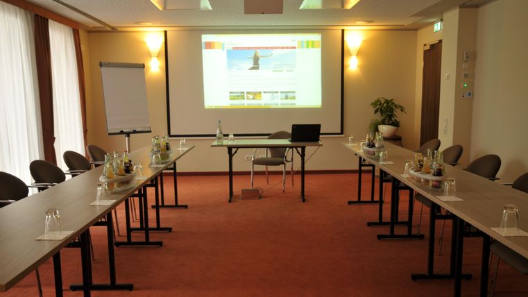 Ein Konferenzraum mit vielen Stühlen und Tischen. An der Wand hängt ein Bildschirm mit einer Präsentation.