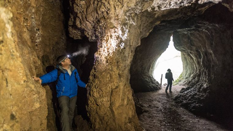 Zwei Personen in einer Höhle, eine mit Stirnlampe, die andere im Hintergrund am Ausgang.