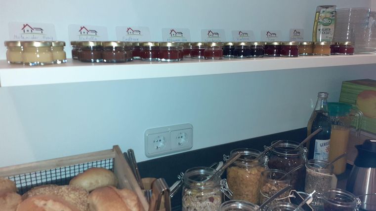 Eine Auswahl an Marmeladen und Honiggläsern steht auf einem Regal. Darunter sind frische Brötchen und verschiedene Vorratsbehälter.