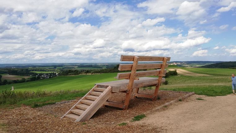 A huge wooden bench with stairs stands in a green landscape under a cloudy sky. A person can be seen on the right edge of the image.