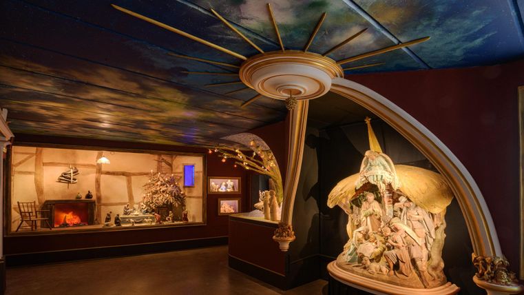 Een kunstzinnig vormgegeven ruimte met indrukwekkende sculpturen en een kleurrijk plafond. In een van de vitrines is een gezellige scène met een open haard te zien.