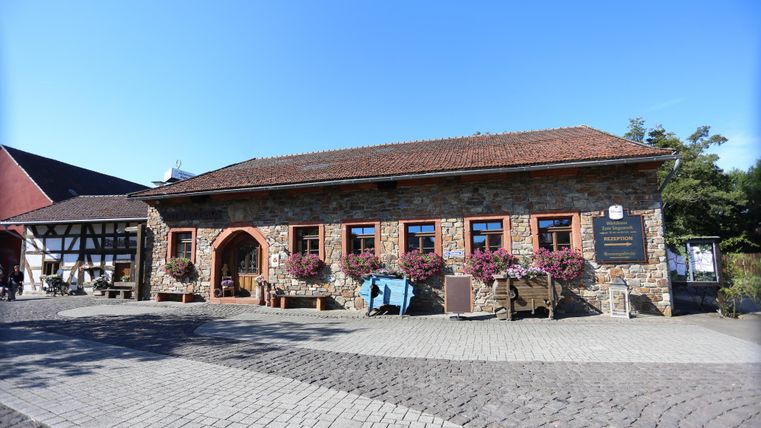 Ein längliches Gebäude mit einer Backsteinfasse, davor ein großer Hof. Neben dem Eingangstor ist ein Schild mit dem Wappen von Rheinland-Pfalz und der Inschrift "Standesamt" angebracht.