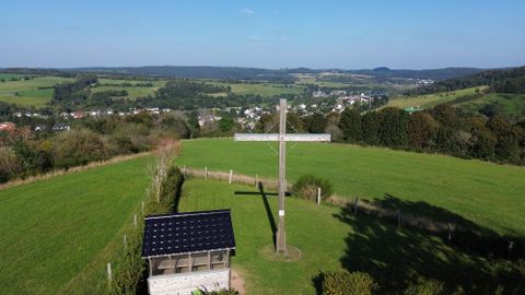 Vogelperspektive auf den Eifelblick auf dem Hasenberg in Stadtkyll mit dem großen Friendenskreuz aus Holz und danebenstehender Schutzhütte. Blick auf das Tal mit Wiesen, Wäldern und einigen Wohnhäusern.