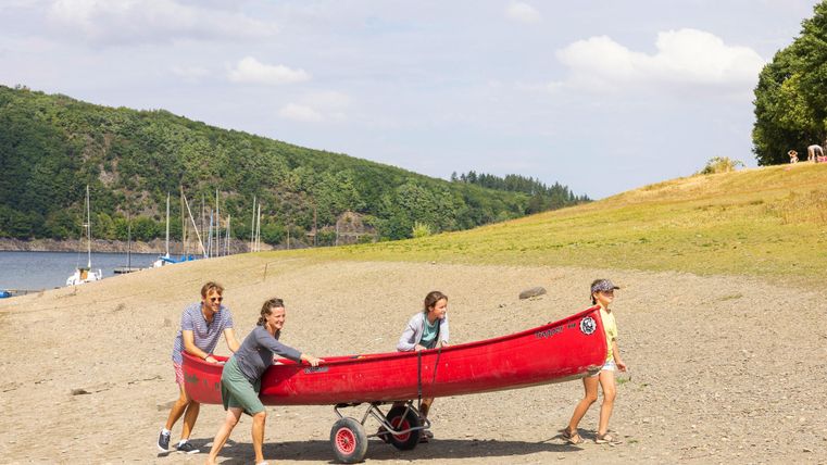 Een groep van vier personen trekt een rode kano naar het strand. Op de achtergrond zijn groene heuvels en een rustig watergebied te zien.