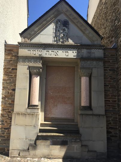 Ein historisches Gebäude mit einem dreieckigen Dach und Säulen. Der Eingang ist umgeben von Steinornamenten und verleiht dem Gebäude einen besonderen Charakter.