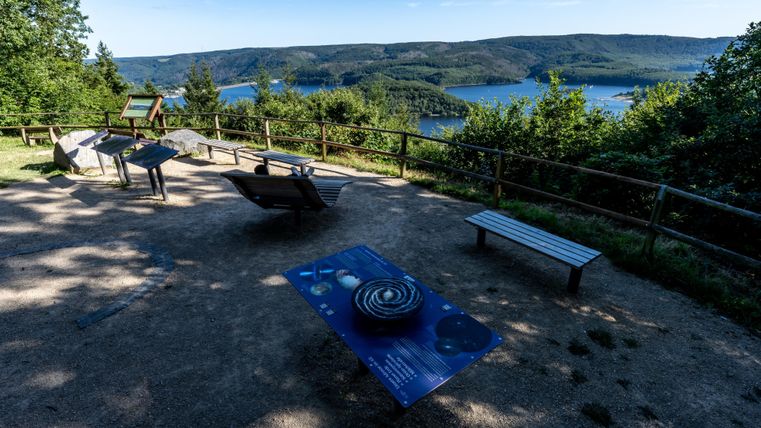 Eine Aussichtsplattform mit einem herrlichen Blick auf einen See und umliegende Hügel. Es gibt mehrere Sitzgelegenheiten und Informationstafeln in der Nähe.