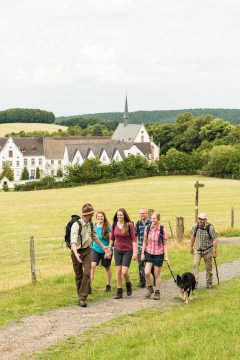 Een groep wandelaars loopt een pad af, vergezeld door een hond. Op de achtergrond is een mooi, oud gebouw en groene velden te zien.