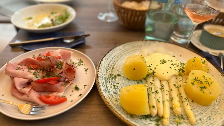 Ein Tisch mit zwei Tellern. Links eine Platte mit Schinken und Tomaten, rechts Spargel und Kartoffeln, garniert mit Kräutern.