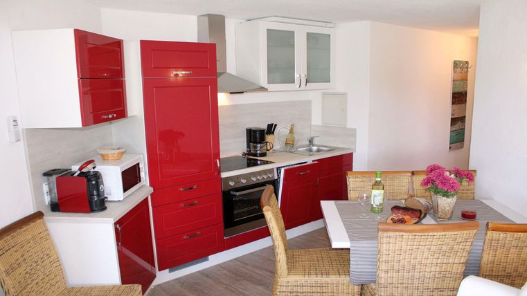 Eine moderne Küche mit roten Schränken und weißen Wänden. Der Essbereich ist gemütlich mit einem Tisch und Stühlen aus Rattan eingerichtet.