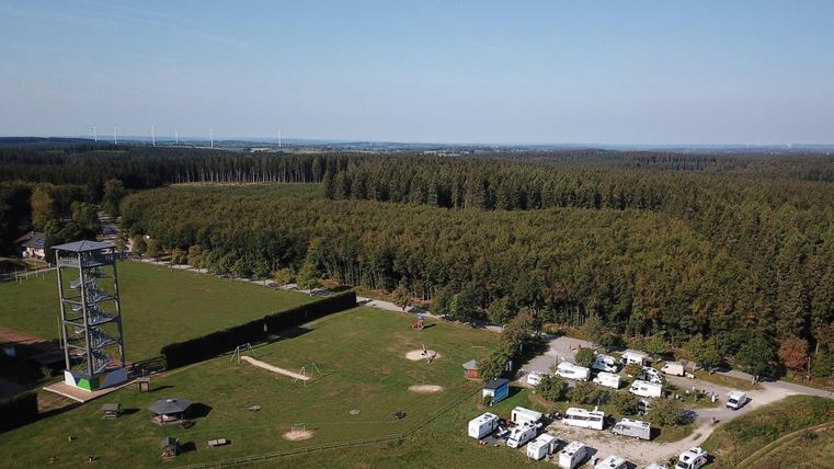 Eine große Wiese mit Wohnmobilen und einem Wald im Hintergrund. Im Vordergrund steht ein Aussichtsturm und ein Spielplatz.