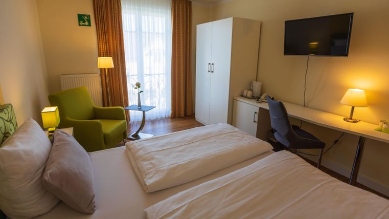 Ein gemütliches Zimmer mit zwei Betten, einem Schreibtisch und einem bequemen Sessel. Große Fenster lassen viel Tageslicht herein und sorgen für eine einladende Atmosphäre.