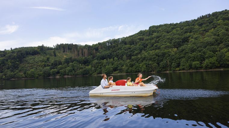 Drei Personen fahren mit einem Tretboot auf einem ruhigen Gewässer. Im Hintergrund sind grüne Hügel und ein klarer Himmel zu sehen.