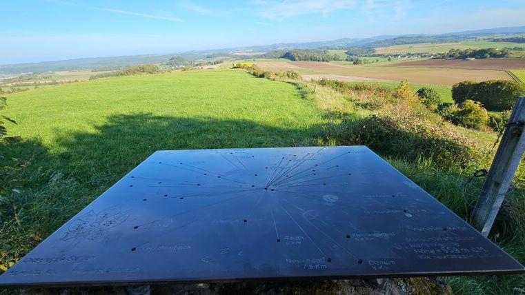 Eine Aussichtsplattform mit einer großen Metalltafel ist zu sehen. Im Hintergrund erstreckt sich eine weite Landschaft mit grünen Wiesen und sanften Hügeln.