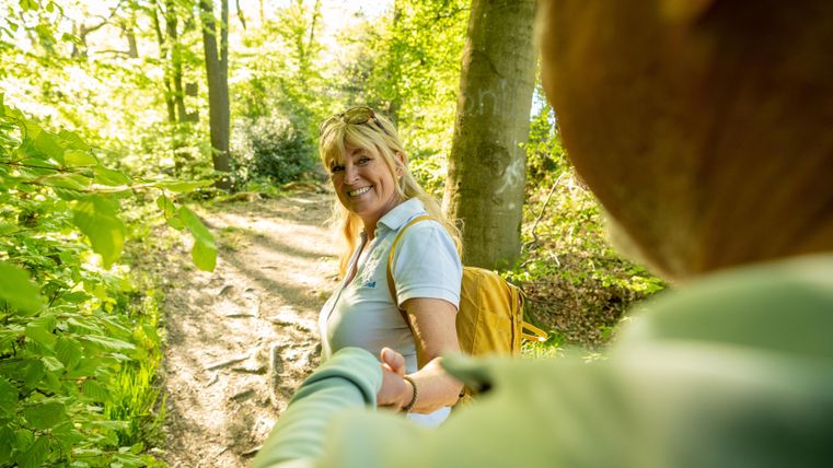A woman smiles on a sunny forest path and holds the hand of another. The surroundings are green and full of trees.