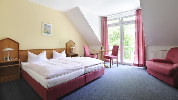 Ein gemütliches Hotelzimmer mit zwei Betten, einem Tisch und einem Sessel. Große Fenster sorgen für viel Tageslicht.