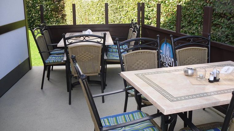 A cozy outdoor area with tables and chairs. The atmosphere is inviting, surrounded by green plants.