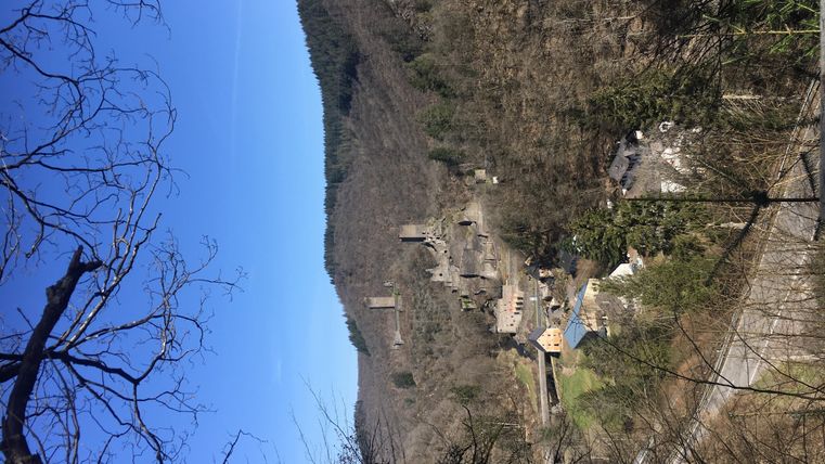 An old ruin in a hilly landscape under a clear blue sky. In the foreground, bare trees are visible.