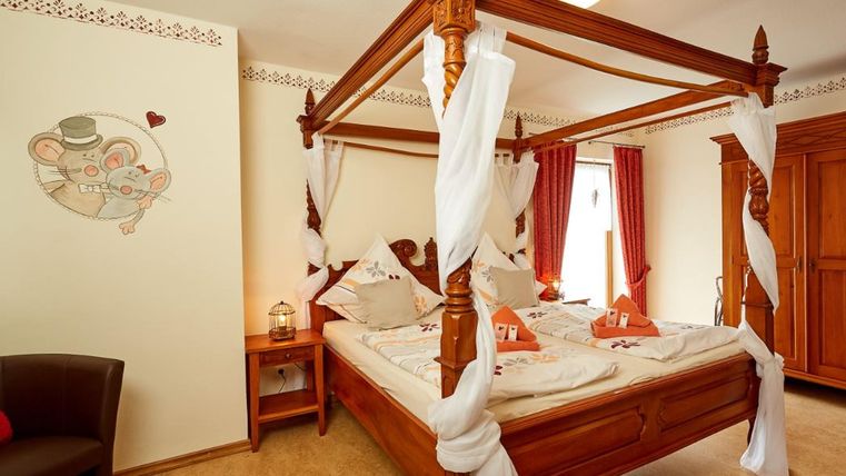 Ein gemütliches Schlafzimmer mit einem Himmelbett aus Holz und dekorativen Kissen. Die Wände sind hell gehalten, und es gibt eine süße Wandmalerei von einer Maus.