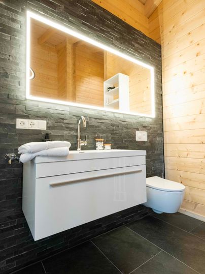 Ein modernes Badezimmer mit einem weißen Waschbecken und einem spiegelnden LED-Spiegel. Die Wände sind in dunklem Stein und hellem Holz gestaltet.