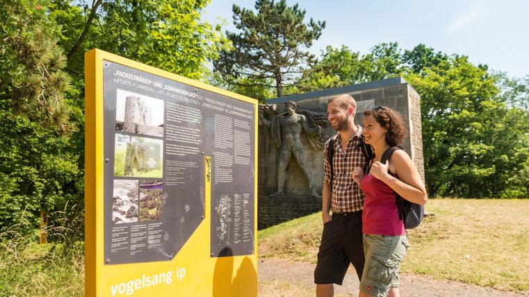 Ein Paar steht vor einem Informationsschild in der Natur. Im Hintergrund sind Bäume und eine Statue sichtbar.