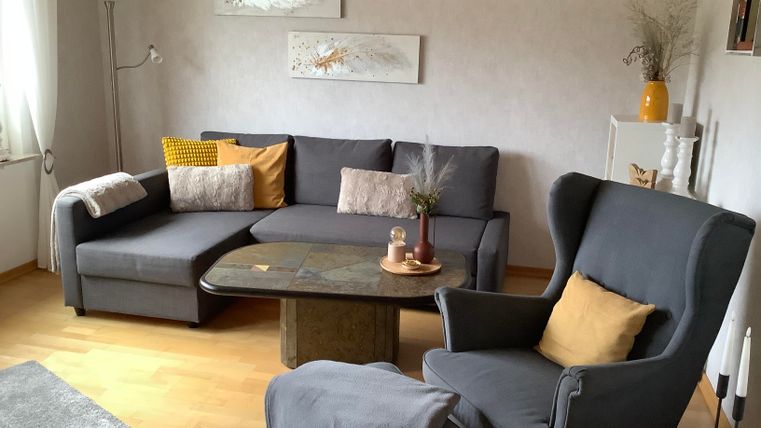 Ein gemütliches Wohnzimmer mit einem grauen Sofa und einem Sessel. Der Tisch in der Mitte hat eine dekorative Kerze und eine entspannte Atmosphäre.