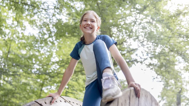 Een vrolijk meisje speelt op een houten element buitenshuis. Op de achtergrond zijn groene bomen te zien.