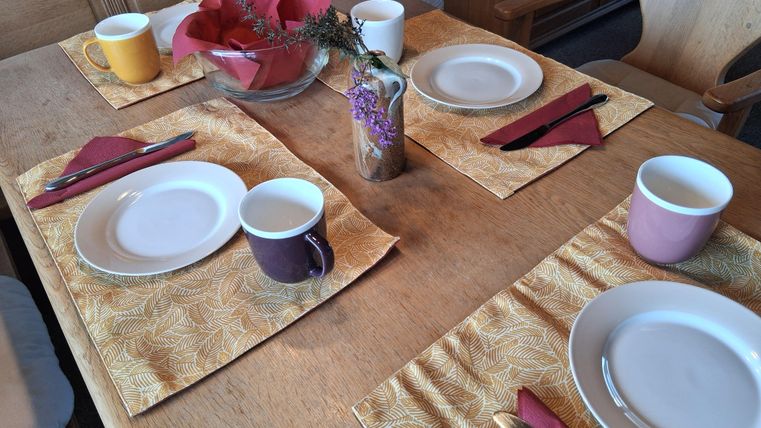 Ein schön gedeckter Tisch mit Tellern, Tassen und bunten Servietten. Die Tischdecke ist gelb mit einem Blattmuster.