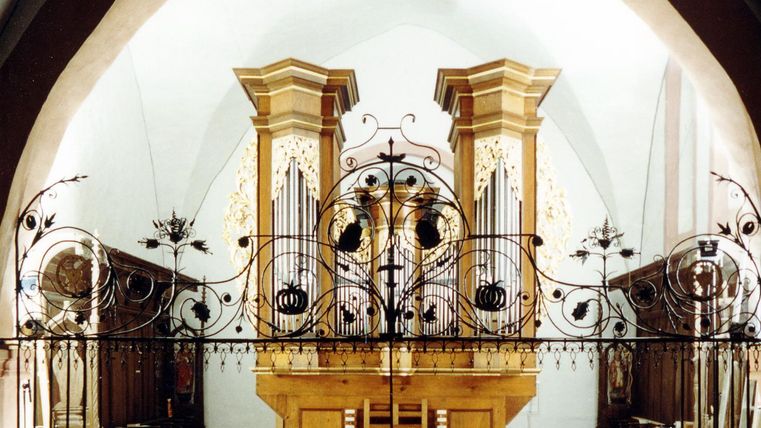 Historische Orgel mit kunstvollem Metallgeländer in einer Kirche, umgeben von Holzverzierungen und goldenen Details.